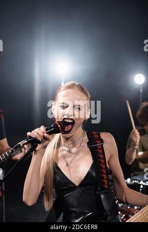 Donna bionda con chitarra basso che urla nel microfono con retroilluminazione e musicisti sfocati sullo sfondo Foto Stock