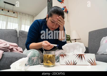 Donna adulta malata seduta sul divano a casa guardando termometro con tè e tovagliolo come malattia di influenza medica concetto di sintomo Foto Stock