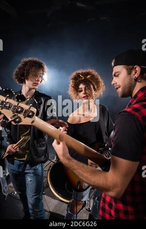 KYIV, UCRAINA - 25 AGOSTO 2020: Uomo curly in piedi vicino a donna e gesturing, mentre si guarda musicista con chitarra elettrica con retroilluminata su nero Foto Stock