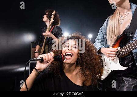KYIV, UCRAINA - 25 AGOSTO 2020: Donna allegra e curly con microfono che canta vicino chitarristi rock band con retroilluminazione su nero Foto Stock