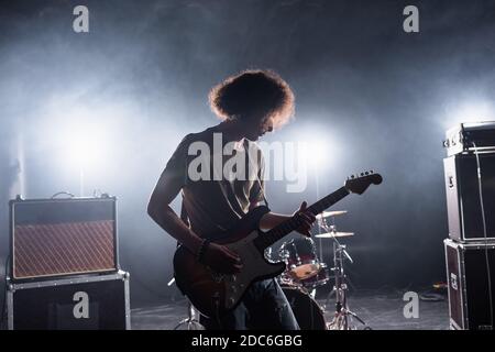 KIEV, UCRAINA - 25 AGOSTO 2020: Curly musicista suonando chitarra basso vicino drum kit e amplificatori combo con retroilluminata su sfondo Foto Stock