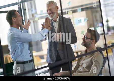 Due uomini d'affari maturi eccitati che danno l'alto cinque l'uno all'altro mentre si ha una riunione con i colleghi nell'ufficio moderno Foto Stock