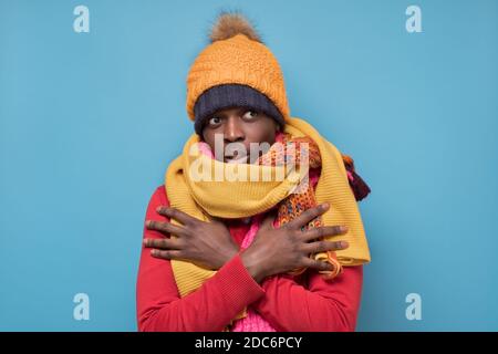 Divertente uomo afroamericano maturo in sciarpa gialla e cappello che si gelano in abiti caldi. Riprese in studio su parete blu. Foto Stock