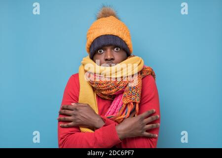 Divertente uomo afroamericano maturo in sciarpa gialla e cappello che si gelano in abiti caldi. Riprese in studio su parete blu. Foto Stock