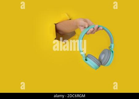 Tenere le cuffie blu a mano su carta gialla strappata. Mano con le cuffie in un foro di carta. Concetto di musica. Foto creativa con spazio per il testo. Min. Alla moda Foto Stock
