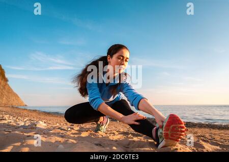 Giovane donna che si allunga sulla spiaggia di sabbia dell'oceano. All'aperto. Sport e stile di vita sano. Vista dal basso. Foto Stock
