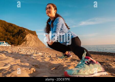 Giovane donna che si allunga sulla spiaggia di sabbia dell'oceano. All'aperto. Sport e stile di vita sano. Vista dal basso. Sneaker da primo piano. Foto Stock