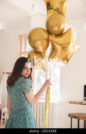 Giovane bella ragazza in abito blu con pois di polka bianco celebra il suo compleanno e gode i palloncini d'oro. Compleanno da solo a casa durante auto-è Foto Stock