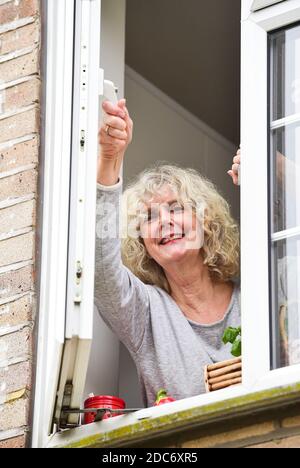 Donna a casa che apre la finestra della cucina per lasciare in fresco Air come consigliato per aiutare a combattere il virus COVID-19 del coronavirus Foto Stock