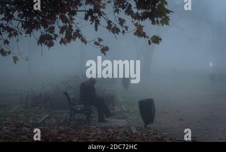 Uomo senza dimora anziano seduto su una panchina nel parco un giorno foggy Foto Stock