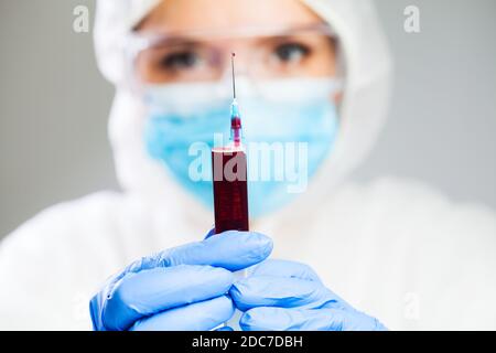 Medico o scienziato di laboratorio in dispositivi di protezione individuale contenenti siringa jab pieno di plasma sanguigno, concentrarsi su goccia singola gocciolando fuori della punta dell'ago Foto Stock