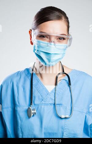 Felice soddisfatta femmina inglese NHS medico indossando maschera protettiva viso & occhiali di sicurezza, occhi sorridenti, studio ritratto isolato su sfondo bianco Foto Stock