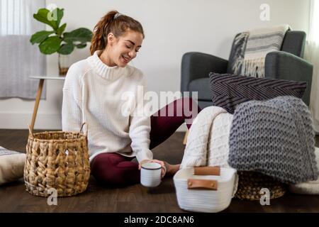 Donna si siede riorganizzando la sua pila di cuscini sul pavimento con la sua bevanda Foto Stock