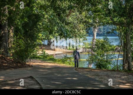 Un uomo che cammina lungo il sentiero attraverso i boschi verso la spiaggia del lago in cerca di un posto andare a pescare mentre si porta il suo placcaggio su un Foto Stock