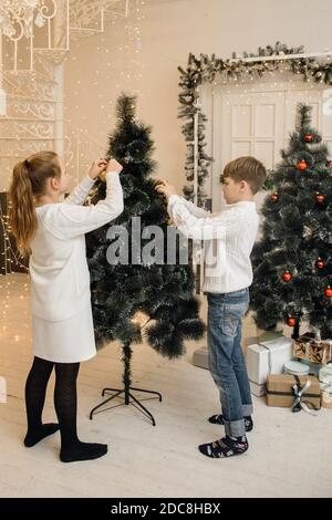 I bambini decorano l'albero di Natale in un interno luminoso. Ragazza e ragazzo, fratello e sorella appendono i giocattoli di Capodanno sui rami di abete rosso. Concetto di Natale e Capodanno per la famiglia. Foto Stock