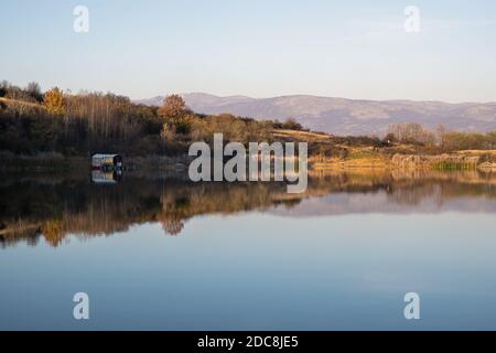 Piccola zattera colorata parcheggiata in un lago calmo, blu argento, riflettente e alberi colorati con colori autunnali Foto Stock