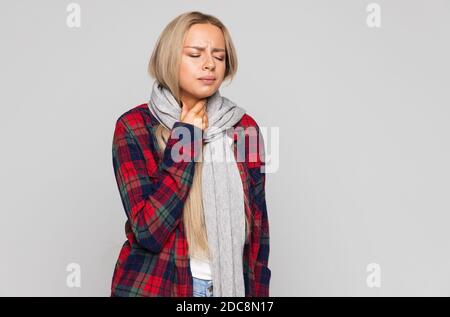 Donna giovane sconvolta in camicia controllata, avvolta in sciarpa con mal di gola, tenendo la mano sul collo. Dolore alla gola, concetto di deglutizione dolorosa. Foto Stock