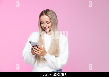 Allegra giovane donna sorridente e utilizzando il telefono cellulare, digitando sms, messaggio, vestito in morbido pullover bianco, isolato su sfondo rosa studio Foto Stock