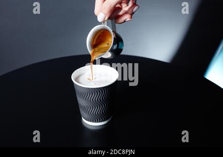 Il barista versa il caffè espresso in una tazza di carta usa e getta su un tavolo di legno nel caffè. Preparazione di una gustosa bevanda al caffè con latte Foto Stock