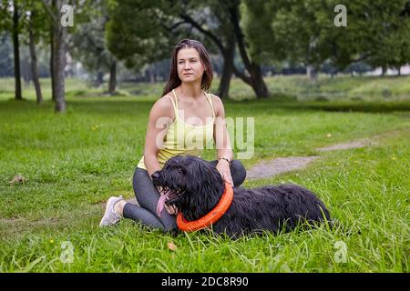 La brida nera si trova sull'erba con la sua proprietaria nel parco pubblico. Ragazza e cane dai capelli lunghi si trovano nel giardino della città. Foto Stock