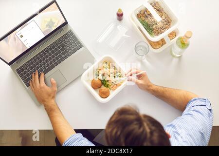 Vista dall'alto dell'uomo che mangia cibo da asporto in contenitori per il pranzo seduti al tavolo bianco di fronte al computer portatile. Foto Stock
