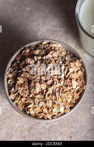 La segale si sfalda in una ciotola con un bicchiere di latte su fondo di pietra. Fiocchi di cereali sani in una ciotola di ceramica e latte da vicino. Foto Stock