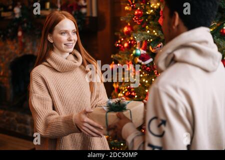 Giovane donna caucasica che dà il regalo di Natale al ragazzo afroamericano sull'albero di natale di sfondo. Foto Stock