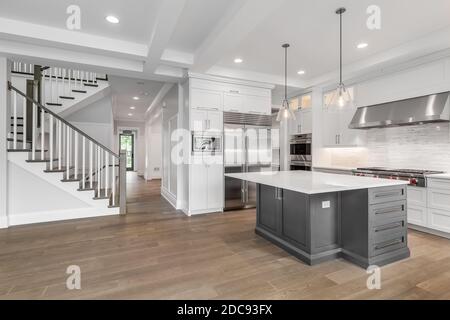 Una casa di lusso con una cucina bianca caratterizzata da un'isola grigia e da elettrodomestici in acciaio inossidabile. Guardando verso una scala e un corridoio per l'ingresso. Foto Stock