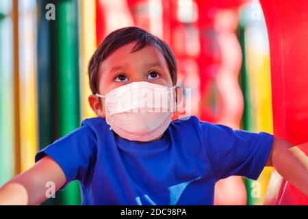 Piccolo ragazzo che si arrampica in un parco giochi per bambini mentre indossa una maschera facciale per proteggere contro i virus in luoghi pubblici. Foto Stock