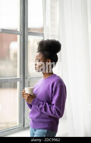 La donna biraciale millenaria afro-americana con orecchini di gioielli alla moda indossano il maglione viola pensando, guardando attraverso la finestra, tenendo il whit Foto Stock