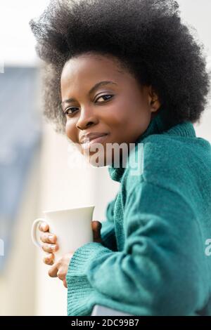 Carina giovane donna afro-americana biraciale godendo di una tazza di tè o caffè, indossare un pullover in maglia verde oversize, guardando la macchina fotografica, in piedi sulla ba Foto Stock