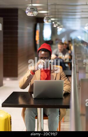 Uomo millenario afro-americano viaggiatore in cappotto beige, cappello rosso seduto al tavolo da caffè nel terminal dell'aeroporto, bere caffè o tè, funziona a distanza sul lapt Foto Stock