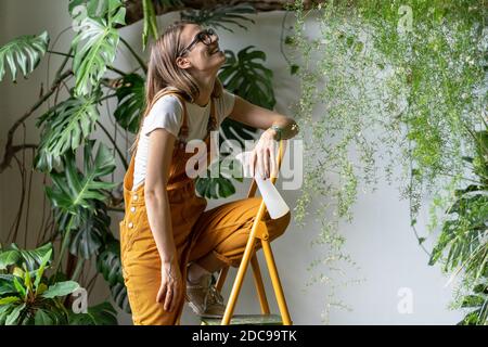 Felice donna giardiniere che tiene pulverizer spray, sorridente, prende una pausa dal lavoro, seduta sulla scala, indossando occhiali e tute arancioni. Verde Foto Stock