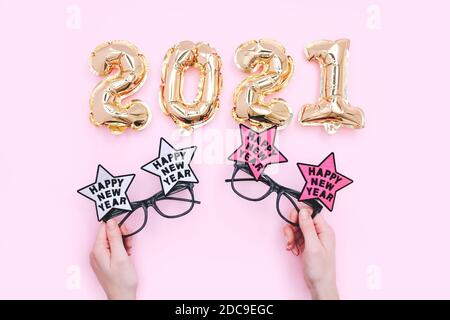 Palloncini in alluminio sotto forma di numeri 2021 e bicchieri di festa in mani femminili. Festa di nuovo anno. Vista dall'alto su sfondo rosa Foto Stock