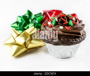 Vista ravvicinata di una cupcake natalizia al cioccolato con glassa di cioccolato e spruzzi rossi, bianchi e verdi. Tre archi di oro, verde, e rosso vento arou Foto Stock