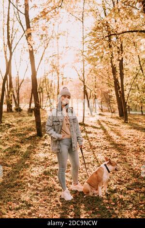 La lunghezza intera di una giovane ragazza allegra adolescente con un cappello in piedi nel parco vicino ad un cane marrone americano Breed Terrier. Passeggiata in autunno nella natura Foto Stock