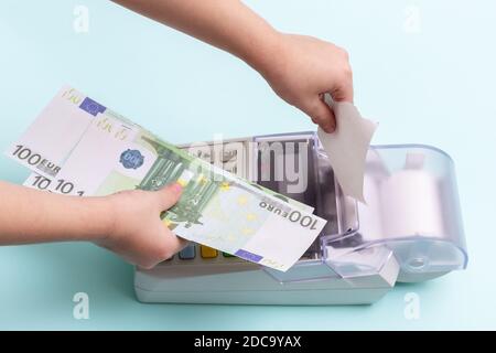 Primo piano di una mano di un bambino che tesse un assegno di cassa in bianco dal terminale e di una mano che tiene diverse centinaia di banconote in euro su sfondo blu, t Foto Stock