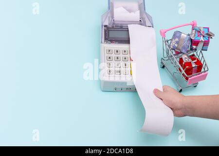 Primo piano della mano di un bambino che tiene in mano la ricevuta di un cassiere in bianco sopra il registratore di cassa accanto a un carrello con confezioni regalo confezionate su uno sfondo blu Foto Stock