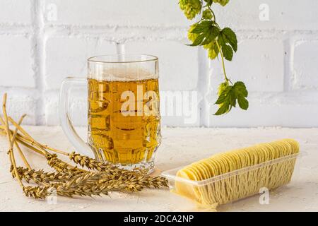 Tazza di vetro di birra con patatine, spighe di orzo e un ramo di luppolo su sfondo bianco. Foto Stock