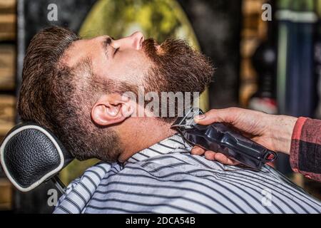 Uomo bearded in barbiere. Uomo in visita hairstylist in barbershop. Barber funziona con un regolabarba. Cliente hipster ottenere haircut. Mani di a. Foto Stock