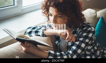 Donna concentrata con capelli ricci sta leggendo un libro mentre seduto a letto vicino alla finestra Foto Stock