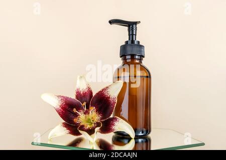 Bottiglia di vetro marrone con pompa di prodotti cosmetici e fiore di giglio su specchio, fondo beige. Natural Organic Spa Beauty concetto cosmetico Mockup. Foto Stock