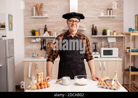 Ritratto dello chef che indossa bonete guardando la fotocamera e sorridendo. Panettiere anziani in cucina uniforme preparare ingredienti di pasticceria su tavola di legno pronti a cucinare pane, torte e pasta gustosi fatti in casa Foto Stock