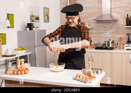 Chef pensionato cracking uova per farina di grano in cucina casa. Cuoco di pasta anziano che coglie l'uovo sulla ciotola di vetro per la ricetta della torta in cucina, mescolando a mano, impastando. Foto Stock