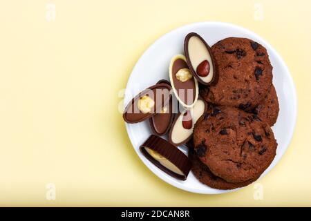 Biscotti e caramelle al cioccolato su un piatto bianco. Vista dall'alto, sfondo giallo Foto Stock