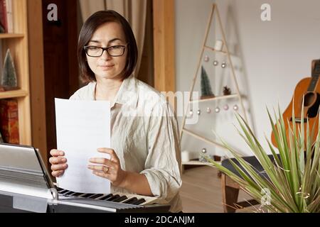 Donna brunetta che ha una chat video con gli amici e suona il pianoforte a casa. Formazione e intrattenimento musicale. Lezioni di musica online. Foto Stock