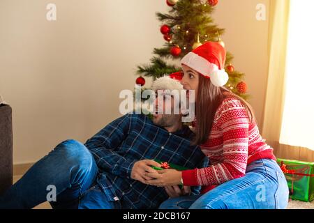 giovane coppia sorprende guardando il lato darsi un regalo con un albero di natale sullo sfondo Foto Stock