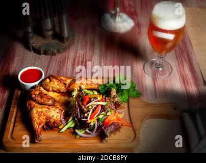 Pollo fritto succoso con verdure ed erbe e un bicchiere di birra leggera sul tavolo nel bar in colori scuri Foto Stock