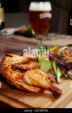 Pollo fritto succoso con verdure ed erbe e un bicchiere di birra leggera sul tavolo nel bar in colori scuri Foto Stock