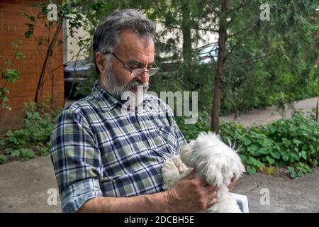 Un uomo sta giocando con il suo piccolo cane maltese. Entrambi godono dell'ombra degli alberi e del giardino dove vivono. Foto Stock